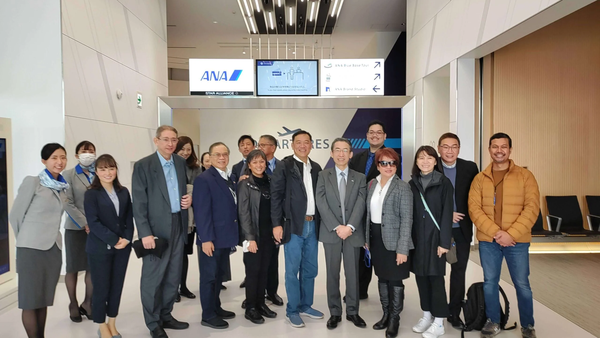 Group photo of diverse people standing in an ANA airport lounge with a sign reading "ANA" and "STAR ALLIANCE".