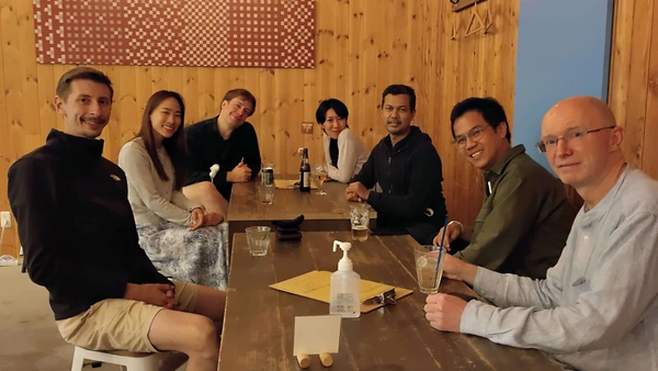 Six people sitting around a wooden table with drinks in a wood-paneled room.