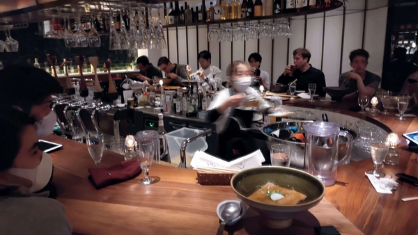 People wearing masks sitting at a curved wooden bar in a restaurant during a tech meetup in Karuizawa.